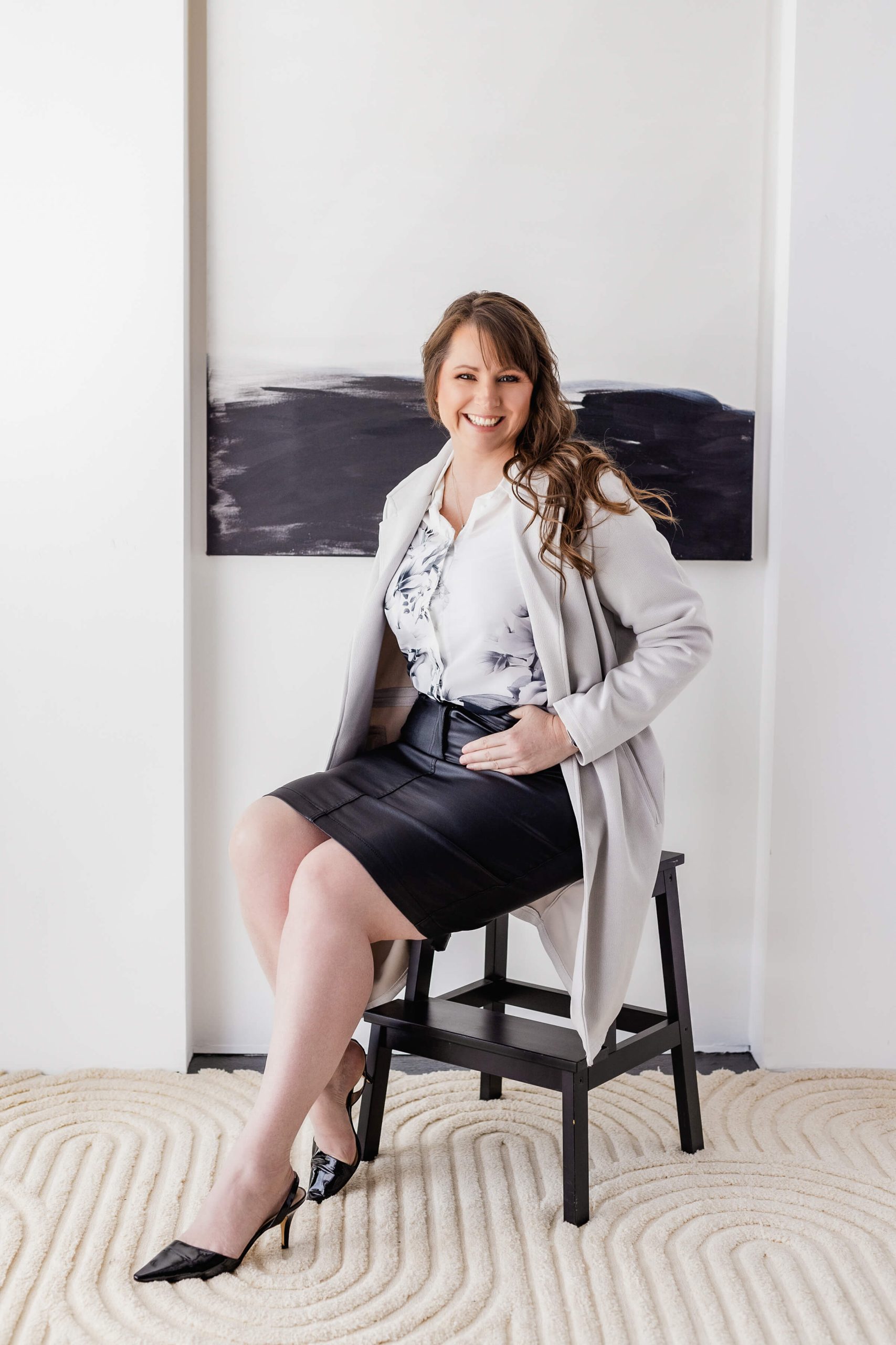 A woman dressed in business attire sits on a stool, smiling. She wears a white blouse, light gray coat, black skirt, and black heels. The background features a black and white abstract painting.