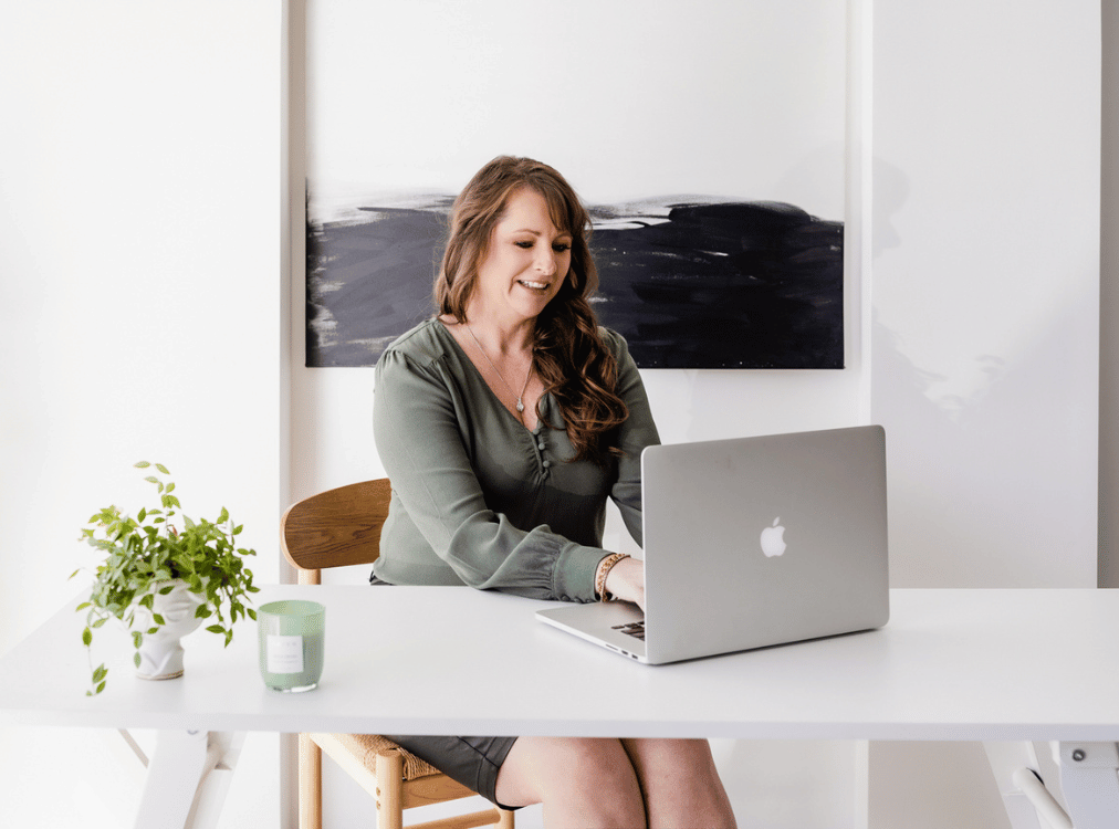 Woman seated at a white desk working on a laptop with a green candle and plant nearby. A black and white abstract painting hangs on the wall behind her. Woman seated at a white desk working on a laptop with a green candle and plant nearby. A black and white abstract painting hangs on the wall behind her.