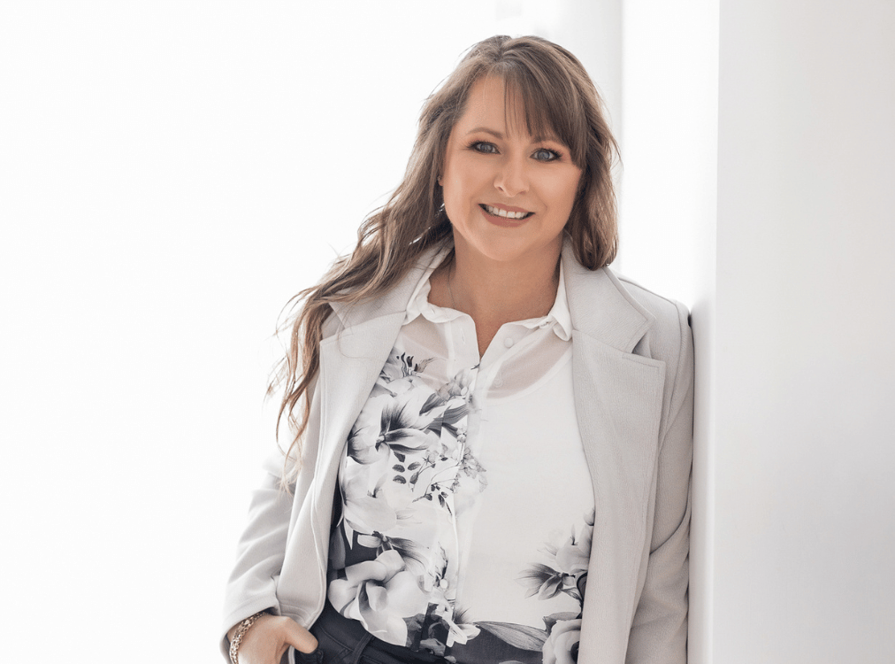 Woman with long brown hair, wearing a floral shirt and light blazer, smiles while leaning against a white wall. Woman with long brown hair, wearing a floral shirt and light blazer, smiles while leaning against a white wall.