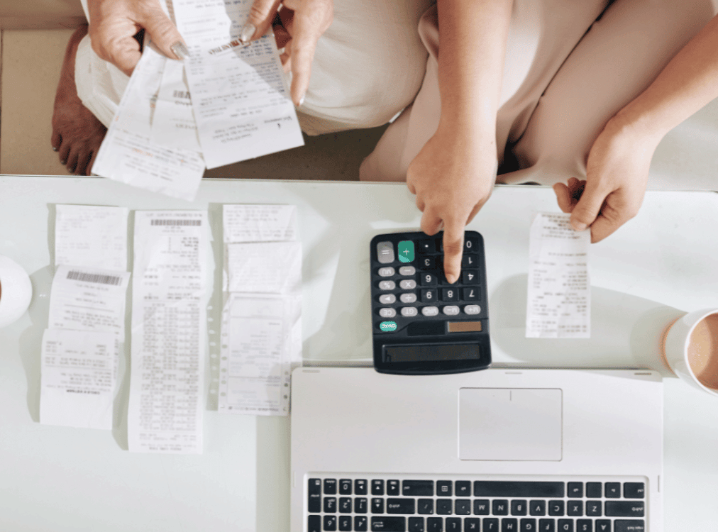 Two people are examining receipts at a table, with a laptop, a calculator, and a cup of coffee also present. Two people are examining receipts at a table, with a laptop, a calculator, and a cup of coffee also present.