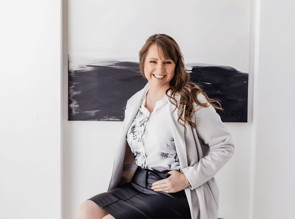 A woman is sitting and smiling, dressed in a light gray blazer, white floral blouse, and black skirt. She has long hair and is posing in front of a black and white abstract painting. A woman is sitting and smiling, dressed in a light gray blazer, white floral blouse, and black skirt. She has long hair and is posing in front of a black and white abstract painting.